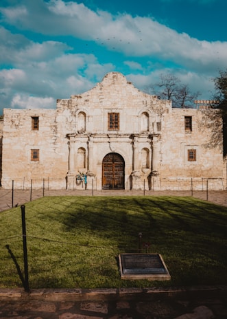 A historic stone building with an ornate facade and arched doorway. The architecture features several small, square windows and aged textures. It is set against a bright blue sky with some fluffy clouds and a small flock of birds. In the foreground, well-maintained green grass is framed by a low rope barrier, and a plaque is embedded in the ground.