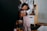 A person in a gray sweater cradles a child, who is wearing pink pajamas with cartoon characters. They are in a room with a wooden floor, surrounded by children's toys and furniture.