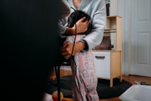 A person in a gray sweater cradles a child, who is wearing pink pajamas with cartoon characters. They are in a room with a wooden floor, surrounded by children's toys and furniture.