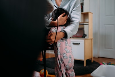 A person in a gray sweater cradles a child, who is wearing pink pajamas with cartoon characters. They are in a room with a wooden floor, surrounded by children's toys and furniture.