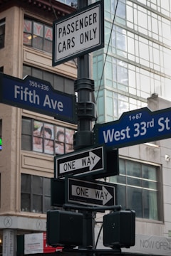 A street sign post located at an urban intersection featuring multiple signs. There are direction signs for 'Fifth Ave' and 'West 33rd St', alongside signage indicating one-way traffic directions. Above these is a sign specifying 'Passenger Cars Only'. The background consists of glass building facades and visible advertisements.