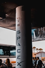 A large vertical structure with 'SpaceX' and 'Falcon 9' written on it is prominently placed. The structure has an American flag on it. Surrounding it are a few people standing and a parked bus. A poster, partially visible, mentions the Kennedy Space Center. The background includes a building and some palm trees.
