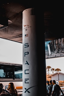 A large vertical structure with 'SpaceX' and 'Falcon 9' written on it is prominently placed. The structure has an American flag on it. Surrounding it are a few people standing and a parked bus. A poster, partially visible, mentions the Kennedy Space Center. The background includes a building and some palm trees.