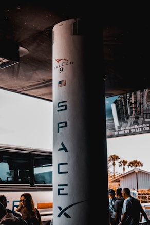 A large vertical structure with 'SpaceX' and 'Falcon 9' written on it is prominently placed. The structure has an American flag on it. Surrounding it are a few people standing and a parked bus. A poster, partially visible, mentions the Kennedy Space Center. The background includes a building and some palm trees.