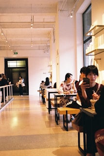 A spacious, modern cafe with high ceilings and warm lighting. People sit at long tables, some engaged in conversation, while others are occupied with their phones. Large windows illuminate the interior, contributing to a cozy atmosphere.