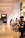 A spacious, modern cafe with high ceilings and warm lighting. People sit at long tables, some engaged in conversation, while others are occupied with their phones. Large windows illuminate the interior, contributing to a cozy atmosphere.