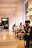 A spacious, modern cafe with high ceilings and warm lighting. People sit at long tables, some engaged in conversation, while others are occupied with their phones. Large windows illuminate the interior, contributing to a cozy atmosphere.