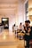 A spacious, modern cafe with high ceilings and warm lighting. People sit at long tables, some engaged in conversation, while others are occupied with their phones. Large windows illuminate the interior, contributing to a cozy atmosphere.