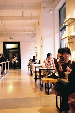 A spacious, modern cafe with high ceilings and warm lighting. People sit at long tables, some engaged in conversation, while others are occupied with their phones. Large windows illuminate the interior, contributing to a cozy atmosphere.