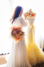 A woman in a white dress holds a colorful bouquet with roses while standing next to a mannequin dressed in a flowing, light yellow gown. The mannequin features a floral-patterned torso. The setting is bright and airy, suggesting a cheerful atmosphere.