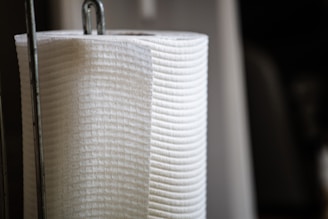 A close-up of a vertically oriented white paper towel roll, resting on a metal holder with a focus on the texture and material of the towel.