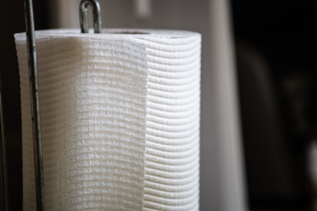 A close-up of a vertically oriented white paper towel roll, resting on a metal holder with a focus on the texture and material of the towel.