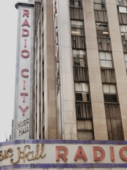 A tall building is partially visible, featuring a vertical neon sign with the words 'Radio City' prominently displayed. Below, another sign reads 'Music Hall' with the text 'Radio City' running horizontally across a marquee. The building exterior is composed of concrete and glass windows.