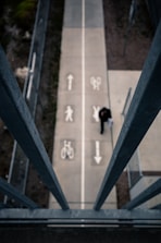 A top-down view of a pedestrian and bicycle path, divided by painted symbols indicating separate lanes for bikes and pedestrians. A person walks along the path, which is flanked by steel railings. The scene is viewed from an elevated perspective, surrounded by patches of greenery and concrete.