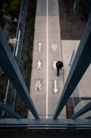 A top-down view of a pedestrian and bicycle path, divided by painted symbols indicating separate lanes for bikes and pedestrians. A person walks along the path, which is flanked by steel railings. The scene is viewed from an elevated perspective, surrounded by patches of greenery and concrete.