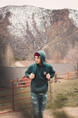 A person wearing a green hoodie and a beanie is walking through a rural area with snow-capped mountains in the background. They have a backpack and are strolling alongside a red fence and some grassy terrain, with houses visible in the distance.