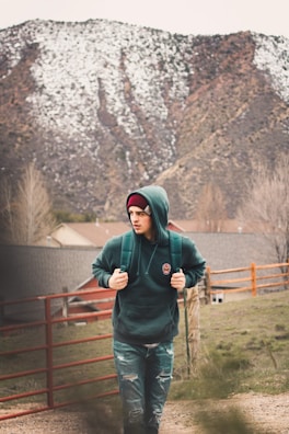 A person wearing a green hoodie and a beanie is walking through a rural area with snow-capped mountains in the background. They have a backpack and are strolling alongside a red fence and some grassy terrain, with houses visible in the distance.