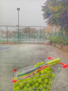 A wire basket filled with numerous tennis balls and a pair of tennis racquets is positioned on an outdoor tennis court. The court is surrounded by a tall chain-link fence. The scene is set on a foggy day with trees in the background and an overcast sky. Visible leaves on the ground suggest late autumn.