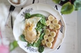 A white plate with ornate black designs holds a breakfast of a fried egg on a bed of fresh spinach leaves and two slices of toast topped with banana slices. The setting includes a blurred background with hints of a coffee cup and partially visible plants.