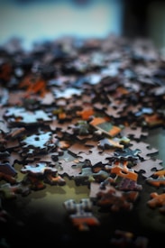A close-up of scattered puzzle pieces on a reflective surface, featuring various shapes and colors. The image is slightly blurred, creating a soft focus effect.