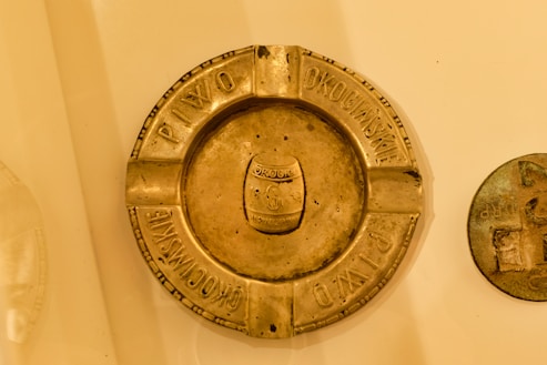 A vintage metallic ashtray with four cigarette rests. The ashtray has an embossed emblem in the center displaying a shield and an inscription. Around the perimeter, text is engraved in a circular fashion. Another round metallic object is visible to the right of the ashtray.