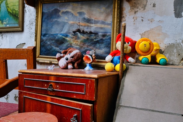 A rustic setting featuring a wooden table with a small drawer, displaying several plush toys. A stuffed dog and a red cartoon character toy sit on top of the table. A colorful toy lion is nearby. In the background, there is an oil painting depicting a ship battling stormy seas, framed in an ornate frame, adding a dramatic touch to the scene. The wall shows signs of peeling paint and wear, enhancing the vintage and weathered ambiance.