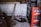 A cluttered storage space featuring stacks of crates filled with empty Coca Cola bottles. A red fire extinguisher is on the floor among scattered wooden planks and debris. The walls are bare and concrete.