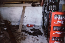 A cluttered storage space featuring stacks of crates filled with empty Coca Cola bottles. A red fire extinguisher is on the floor among scattered wooden planks and debris. The walls are bare and concrete.