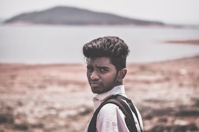 A young man with short black hair and a light beard is looking over his shoulder towards the camera while wearing a backpack. He stands on a dry, rocky landscape with a blurred background of a large body of water and a hilly landmass in the distance.