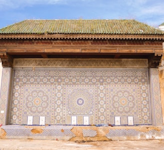 An ornate wall decoration features intricate geometric patterns with a mosaic of colorful tiles, predominantly in shades of blue, yellow, and white. The design includes circular and floral motifs, arranged symmetrically across the facade. A green-tiled roof with a wooden overhang complements the detailed artistic work below.