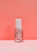 A bottle of nail polish with a white cap and a clear container showcasing pink glitter suspended in the liquid. The background is a soft pink color that complements the bottle's contents.