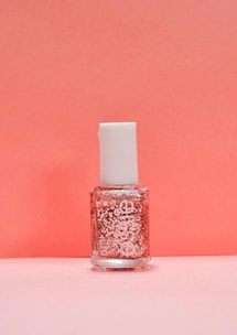 A bottle of nail polish with a white cap and a clear container showcasing pink glitter suspended in the liquid. The background is a soft pink color that complements the bottle's contents.