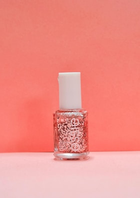 A bottle of nail polish with a white cap and a clear container showcasing pink glitter suspended in the liquid. The background is a soft pink color that complements the bottle's contents.