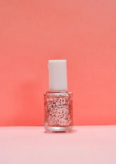 A bottle of nail polish with a white cap and a clear container showcasing pink glitter suspended in the liquid. The background is a soft pink color that complements the bottle's contents.