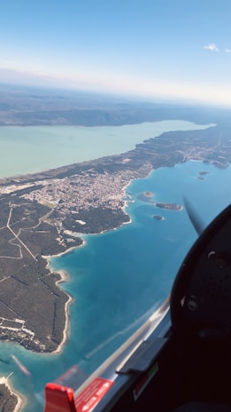 An aerial view of a coastline featuring a sprawling landscape with a mix of land and water formations. The image includes a vast body of water with a turquoise hue juxtaposed against green, richly forested land, and a small town visible along the coast. The sky is clear with a few clouds, and a partial view from the interior of an aircraft with a red and black cockpit is present in the foreground.