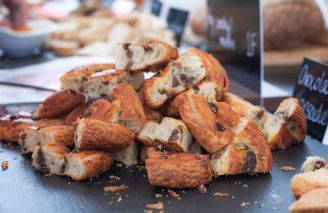 A stack of sliced pastries with golden-brown flaky crusts, filled with what appears to be raisins or chocolate chips. The pastries are arranged on a dark surface, and there are blurred signs in the background indicating a bakery setting.