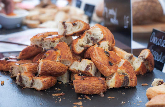 A stack of sliced pastries with golden-brown flaky crusts, filled with what appears to be raisins or chocolate chips. The pastries are arranged on a dark surface, and there are blurred signs in the background indicating a bakery setting.