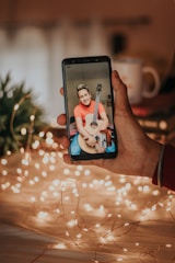 A hand holds a smartphone displaying a person with a guitar. The background features warm, glowing fairy lights and some blurred out objects, creating a cozy atmosphere.