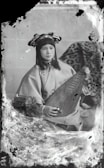 A person wearing traditional attire is seated while holding a stringed musical instrument. The background features a patterned fabric draped over an object. The overall appearance is vintage, with visible signs of age such as scratches and discoloration on the photograph.