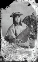 A person wearing traditional attire is seated while holding a stringed musical instrument. The background features a patterned fabric draped over an object. The overall appearance is vintage, with visible signs of age such as scratches and discoloration on the photograph.