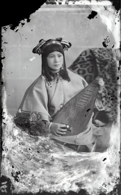 A person wearing traditional attire is seated while holding a stringed musical instrument. The background features a patterned fabric draped over an object. The overall appearance is vintage, with visible signs of age such as scratches and discoloration on the photograph.