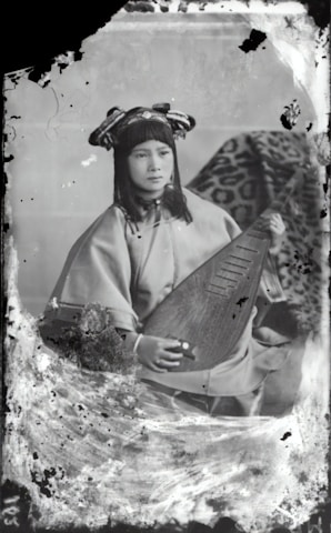 A person wearing traditional attire is seated while holding a stringed musical instrument. The background features a patterned fabric draped over an object. The overall appearance is vintage, with visible signs of age such as scratches and discoloration on the photograph.