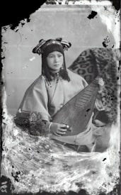 A person wearing traditional attire is seated while holding a stringed musical instrument. The background features a patterned fabric draped over an object. The overall appearance is vintage, with visible signs of age such as scratches and discoloration on the photograph.