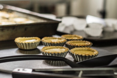 Several small, unbaked pastry shells are arranged on a dark countertop with metal baking trays in the background. The scene suggests a kitchen or baking environment.