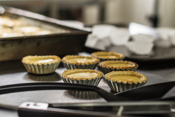 Several small, unbaked pastry shells are arranged on a dark countertop with metal baking trays in the background. The scene suggests a kitchen or baking environment.