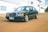 A classic black sedan with a prominent grille parked on a dirt road in front of an industrial building. The car has a sleek design with silver alloy wheels and a distinct license plate. The background features large windows and air conditioning units mounted on the building.