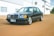 A classic black sedan with a prominent grille parked on a dirt road in front of an industrial building. The car has a sleek design with silver alloy wheels and a distinct license plate. The background features large windows and air conditioning units mounted on the building.
