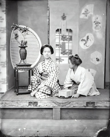Two individuals dressed in traditional Japanese clothing are seated on a raised platform. One person is looking slightly to the side, wearing a checkered patterned kimono. The second person, to their right, is wearing a plain kimono and appears to be holding a small box. Behind them is a decorative screen with floral and geometric designs, and to the left, there is a table with a vase containing branches.
