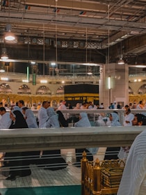 A large group of people, many dressed in traditional Islamic attire, are walking and gathering near a significant religious structure surrounded by elaborate architecture and lighting. The environment appears to be a holy site with a mix of modern and traditional elements.