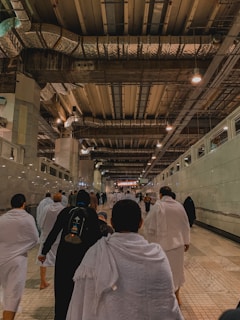 A group of people wearing white garments walk through a large, spacious corridor under a high industrial-style ceiling with exposed ducts and lights. The floor is tiled, and the walls are lined with glass panels. The environment suggests a peaceful and solemn atmosphere as the individuals proceed forward.
